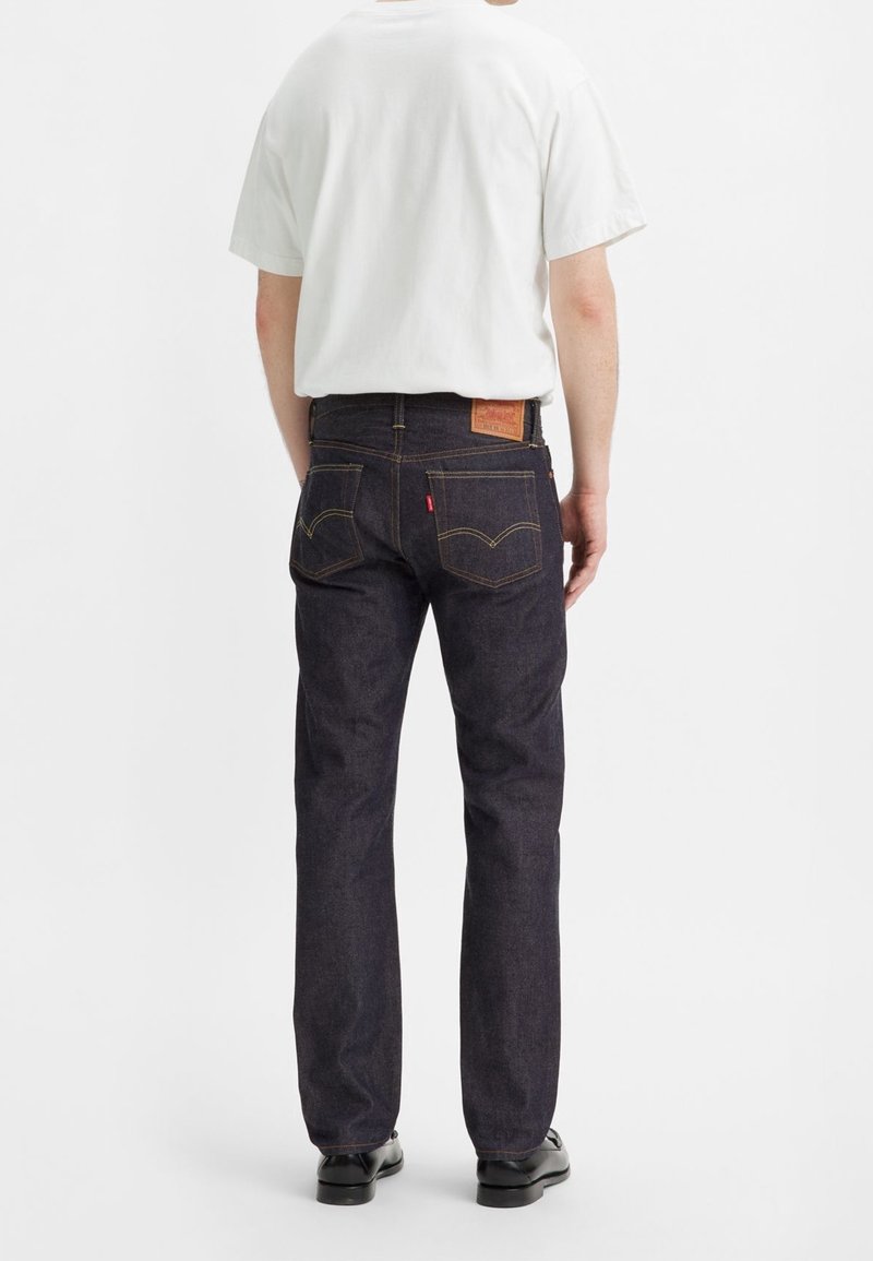 Jean en denim foncé à coupe droite, avec des coutures jaunes, un patch en cuir à l'arrière et une étiquette rouge sur la poche droite.