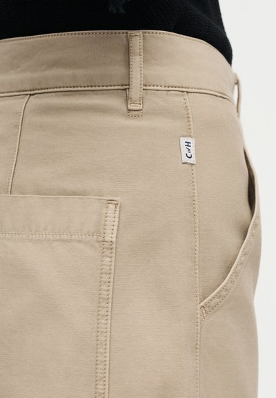 Κοντινό πλάνο μπεζ παντελονιού με ορατή πίσω τσέπη, θηλιά για ζώνη, πλαϊνή τσέπη και μικρή υφασμάτινη ετικέτα με την επιγραφή "CofH".