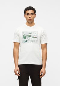 Νέος άνδρας με σγουρά μαλλιά φοράει λευκό T-shirt με σχεδιασμένο πράσινο κροκόδειλο και το κείμενο "LACOSTE" στο μπροστινό μέρος, συνδυασμένο με μαύρο παντελόνι.