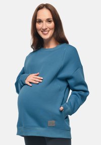 Felpa premaman blu con maniche lunghe, collo rotondo, tasche laterali e tessuto testurizzato. Presenta un'etichetta con logo all'orlo.