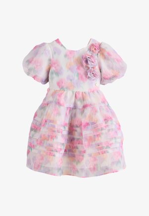 Niña joven de pie con las manos a los lados, vestida con un vestido floral pastel con mangas abullonadas y zapatos Mary Jane dorados.