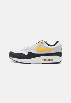 Biało-szary sneaker z czarną podeszwą, żółwym logo Nike po boku, białymi sznurowadłami i widoczną poduszką powietrzną w podeszwie, pokazany z boku.