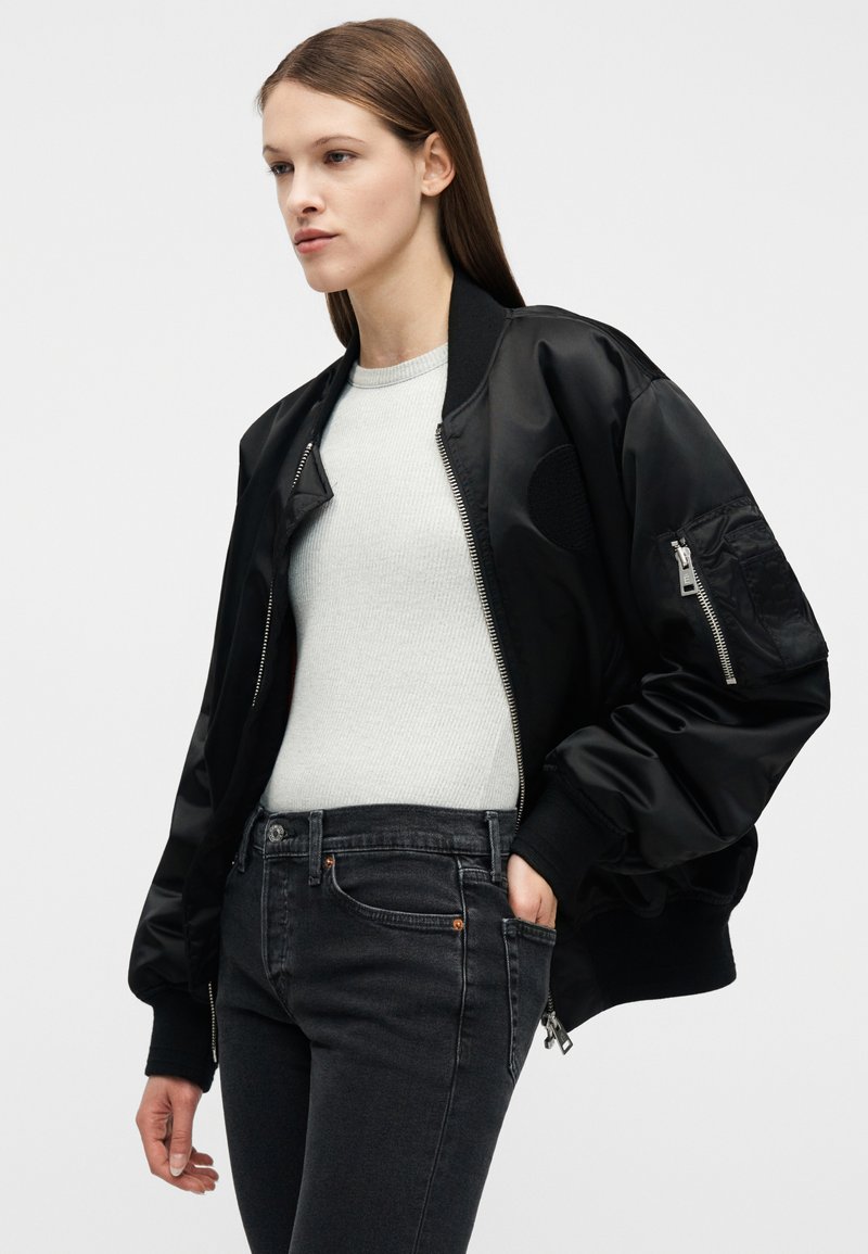 Young woman wearing a black bomber jacket over a light gray ribbed top and dark gray jeans, standing with one hand in pocket.