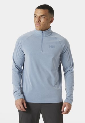 Hombre con sudadera de cremallera corta azul claro con logo de Helly Hansen, de pie y mirando hacia un lado contra un fondo liso.
