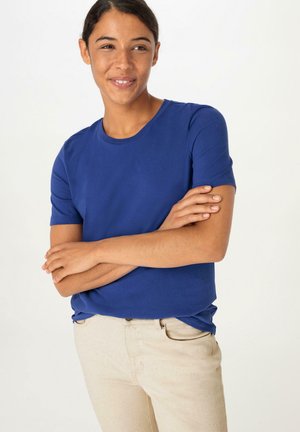 T-shirt in cotone blu con maniche corte e scollatura rotonda, indossato con pantaloni beige chiaro, che mostrano una texture liscia e un design semplice.