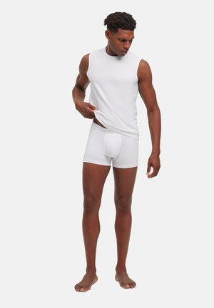 Mann in weißem ärmellosem Shirt und weißen Boxershorts hebt das Shirt leicht an und steht barfuß vor einem schlichten Hintergrund.