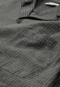 Chemise grise texturée avec un motif en vagues subtil, dotée d'un col, d'un bouton unique et d'une poche poitrine. Le tissu a une finition douce et légèrement côtelée.