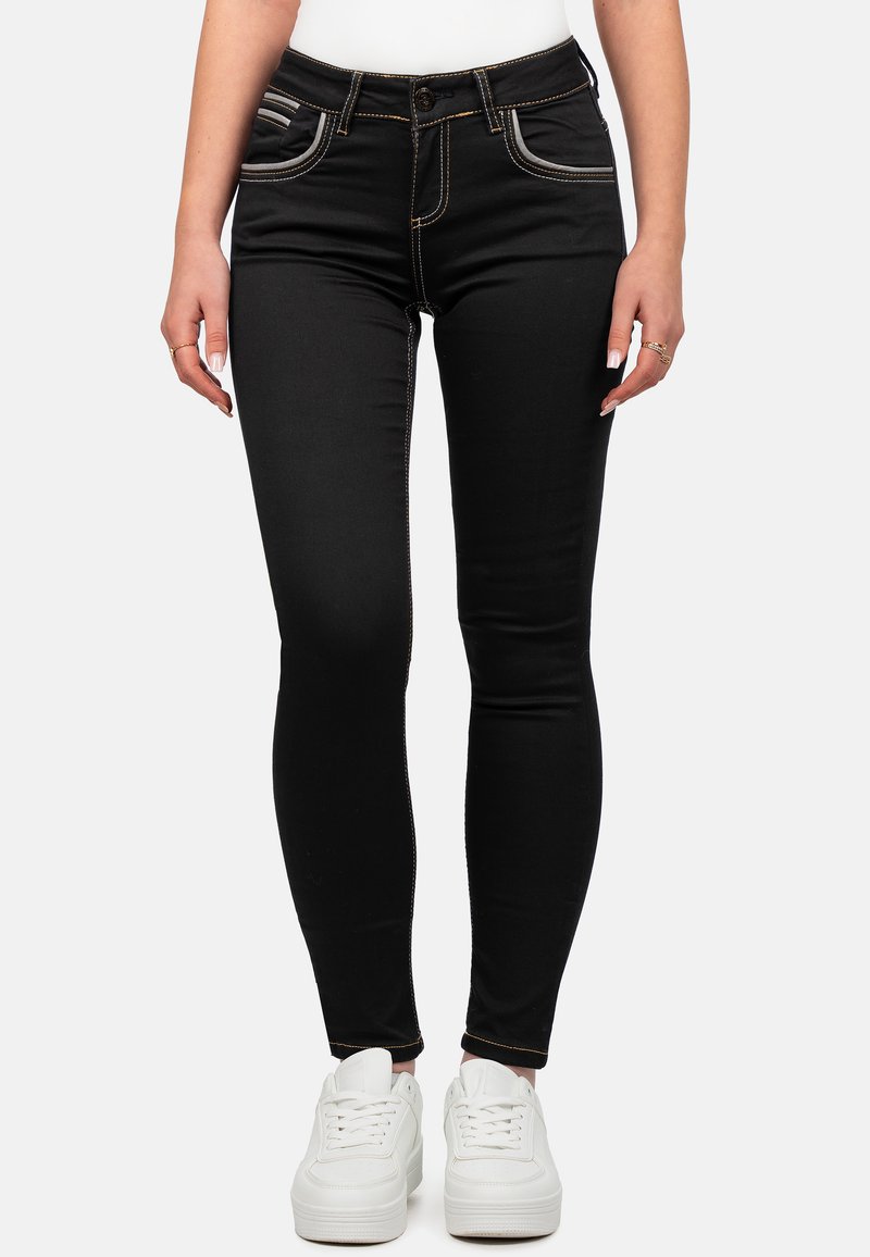 Zwarte skinny jeans met contrasterende gele stiksels, voorzakken en een getailleerd ontwerp, gecombineerd met witte sneakers.