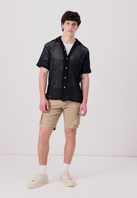 Camisa negra de manga corta con un patrón en zigzag y cierre de botón, combinada con pantalones cortos de cargo beiges y zapatillas beiges. Calcetines blancos visibles.