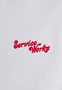 Camiseta de algodón blanca con un logo rojo prominentemente bordado y letras estilizadas que dicen "Service Works".
