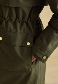 Oliwkowa, wodoodporna kurtka z cinchowanym pasem, zapięciami na zatrzaski oraz przednią kieszenią z wytłoczonym detalem logo.