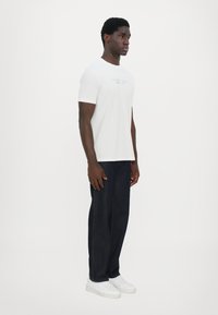 Hvit kortermet t-skjorte med et enkelt tekstlogo på brystet, kombinert med mørk denimjeans og hvite sneakers. Løs passform, glatt tekstur.