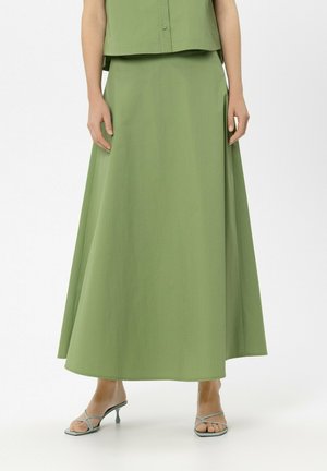 Frau trägt einen langen, grünen A-Linien-Rock in Kombination mit grauen, riemchenbesetzten Absatzsandalen und steht vor einem schlichten weißen Hintergrund.