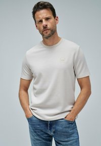 Hombre vistiendo una camiseta de manga corta texturizada color beige claro con un pequeño logo en el pecho y jeans azules, de pie con las manos en los bolsillos frente a un fondo sencillo.