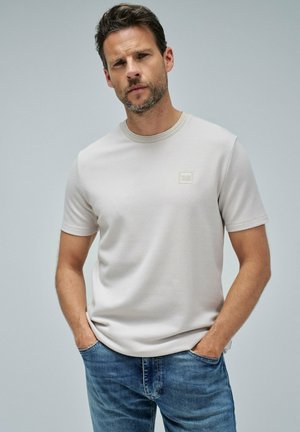 Hombre vistiendo una camiseta de manga corta texturizada color beige claro con un pequeño logo en el pecho y jeans azules, de pie con las manos en los bolsillos frente a un fondo sencillo.