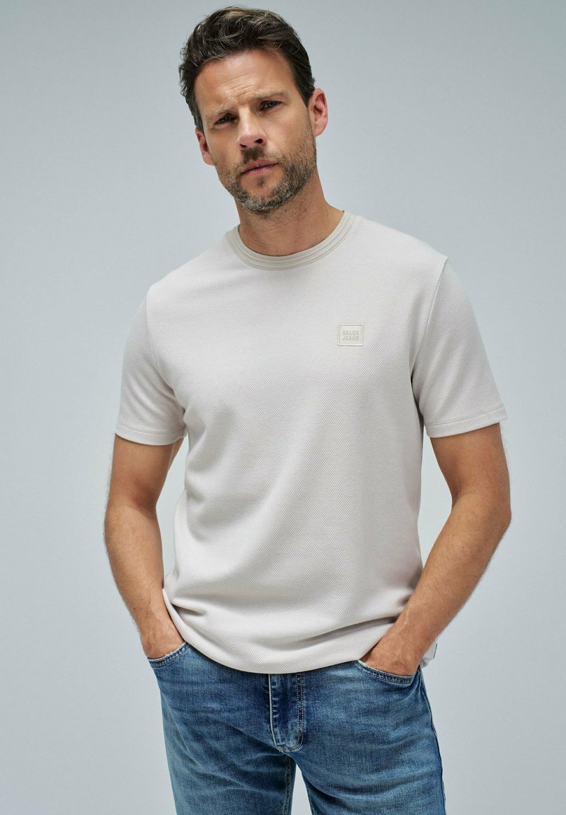 Hombre vistiendo una camiseta de manga corta texturizada color beige claro con un pequeño logo en el pecho y jeans azules, de pie con las manos en los bolsillos frente a un fondo sencillo.