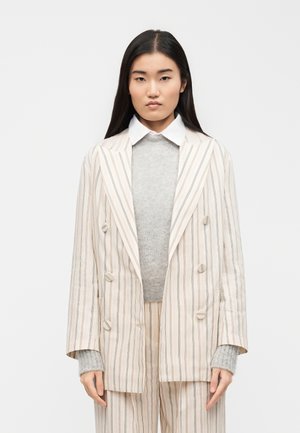 Une femme aux cheveux lisses et foncés porte un blazer crème à rayures fines à double boutonnage par-dessus un pull gris et une chemise blanche à col.