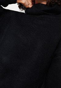 Een persoon met krullend haar die zijn/haar nek aanraakt, gekleed in een dikke zwart geribbelde trui met een hoge kraag.