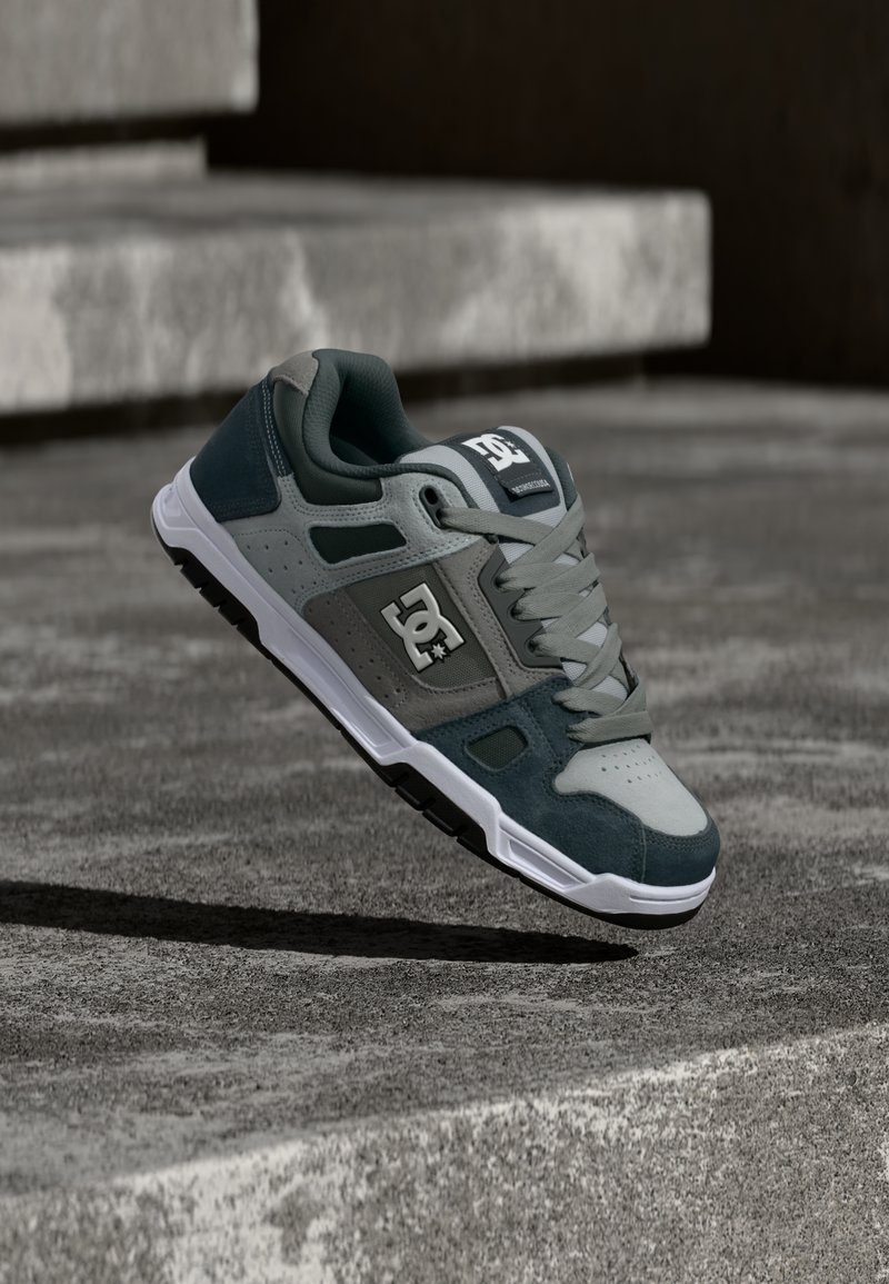 Γκρι και σκούρο πράσινο παπούτσι σκεϊτ DC με λευκή σόλα και λογότυπο, αιωρούμενο πάνω από σκάλες από σκυρόδεμα σε υπαίθριο χώρο.