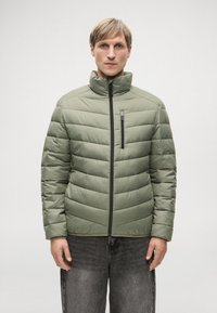 Ελαιόχρωμο αδιάβροχο μπουφάν με ψηλό γιακά, που διαθέτει μπροστινό φερμουάρ και οριζόντια ραφές. Φορεμένο πάνω από σκούρα τζιν.