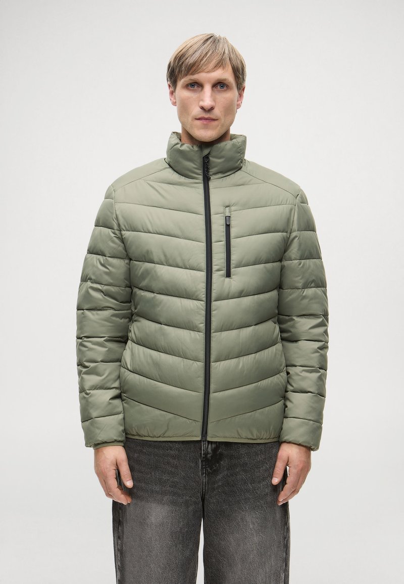 Ελαιόχρωμο αδιάβροχο μπουφάν με ψηλό γιακά, που διαθέτει μπροστινό φερμουάρ και οριζόντια ραφές. Φορεμένο πάνω από σκούρα τζιν.