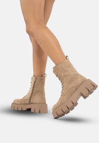 Botas de tobillo de ante en color marrón claro con suela de plataforma gruesa, cordones en la parte delantera y cremallera en el lateral para poner y quitar fácilmente. Acabado texturizado y construcción resistente.