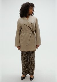 Smėlio spalvos striukė su apvyniojimo stiliumi, plačiomis atlapomis ir diržu, dera su rudo ir juodo dryžuoto zebro rašto kelnėmis bei juodais bateliais.