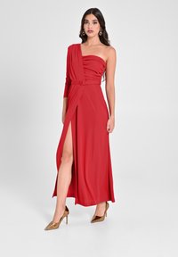 Rotes, einseitig tragendes Kleid mit gerafftem Stoff und einem Seiten-Schlitz, ausgestattet mit einem taillierten Gürtel. Das Model trägt goldene spitze Absätze. Glatte Textur.