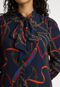 Sieviete tērpusies tumši zilā blūzē ar kakla izgriezumu ar loku, rotātā ar brūnām pinuma auklām, sarkaniem un zilajiem siksnām un zelta ķēžu rakstiem.