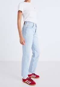 Vit t-shirt med en liten röd logotyp, ljusblå jeans med avslappnad passform, tillsammans med r öda och vita sneakers med mörka sulor.