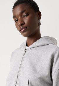 Felpa grigia con zip, tasca frontale, texture morbida e cappuccio regolabile. Presenta una zip argentata e dettagli di cucitura discreti.