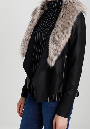 Žena v černé bundě se širokým umělým kožešinovým límcem, černém roláku s tenkými pruhy a modrých džínách, stojí před jednobarevným pozadím.