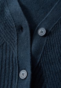 Marinblå stickad kofta med ribbad textur, prydd med runda grå knappar. Nära vy framhäver sömmar och tygets mjukhet.