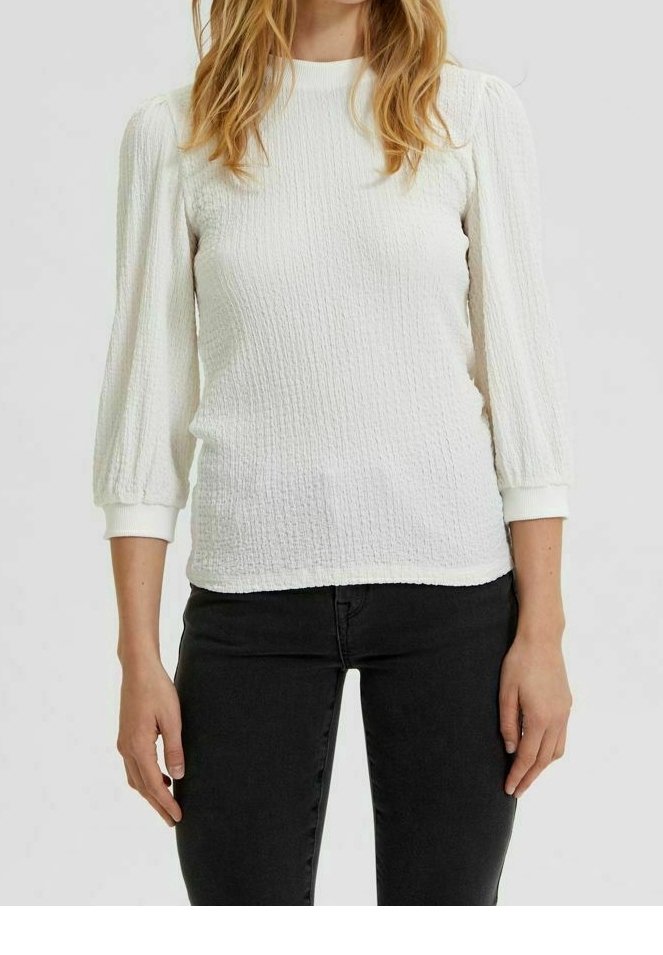 Top bianco testurizzato con scollo a girocollo e maniche a sbuffo a tre quarti, abbinato a jeans neri aderenti. Colore solido, design casual.