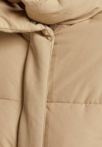 Giacca imbottita beige con una texture liscia, caratterizzata da un collo alto, chiusura con bottoni e cuciture visibili lungo le cuciture.