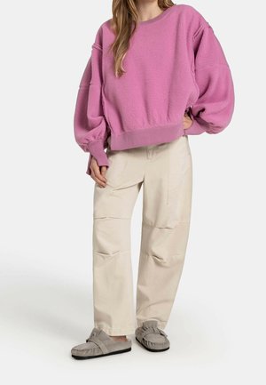 Frau trägt einen lockeren rosa Fleecepullover, beige weite Hosen und graue Mokassin-Slipper und steht mit einer Hand in der Hüfte.