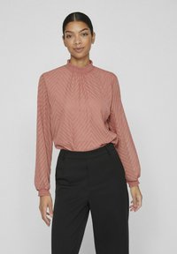 Rosa blus med zigzag-mönster och hög smockad krage samt långa, sydda ärmar. Matchas med svarta skräddarsydda byxor.