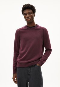 Pull en maille violet avec un col rond côtelé, des coutures complexes sur les épaules et des manches longues, associé à un pantalon gris foncé.