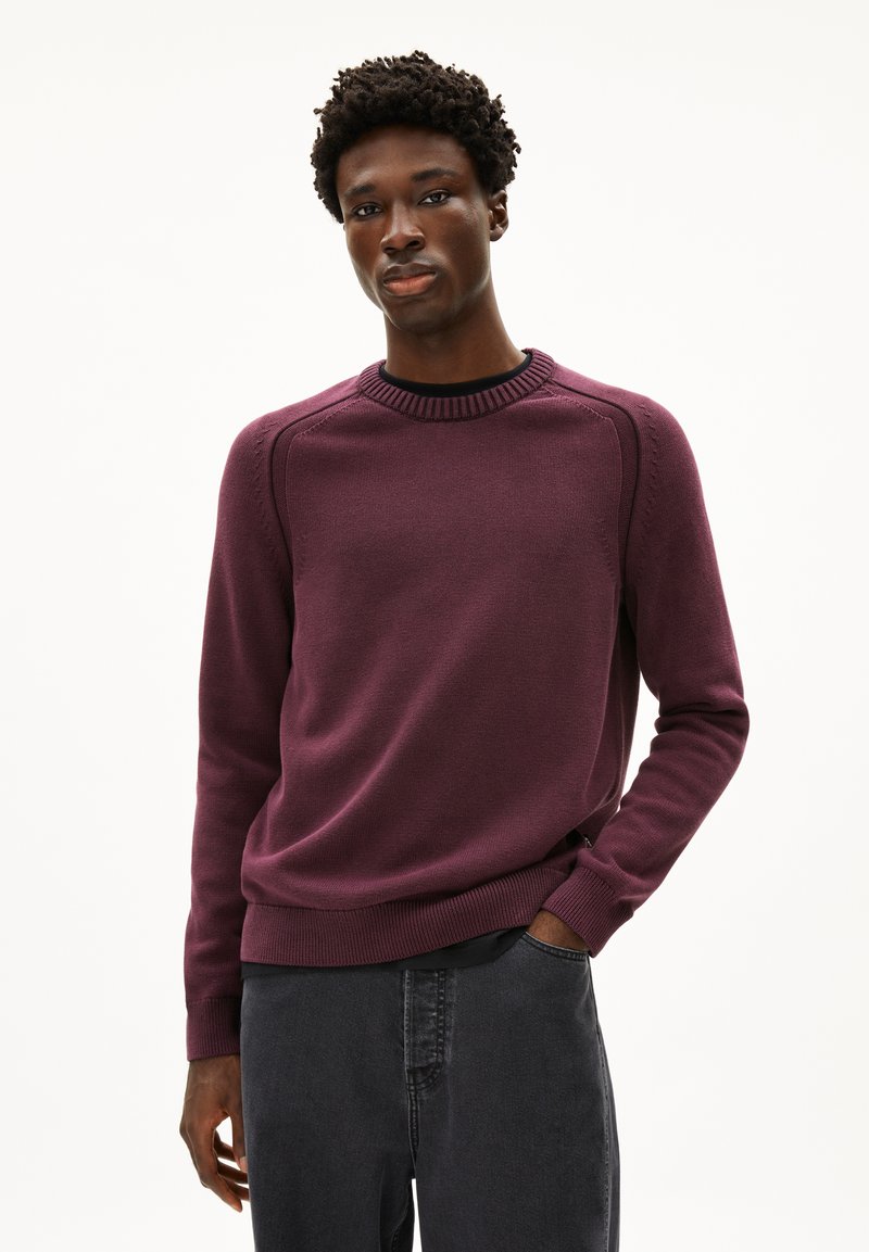 Pull en maille violet avec un col rond côtelé, des coutures complexes sur les épaules et des manches longues, associé à un pantalon gris foncé.