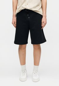 Hombre con pantalones cortos negros hasta la rodilla con cordón, camisa beige, calcetines blancos y zapatillas blancas, de pie contra un fondo liso.