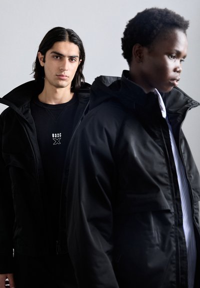 To modeller iført sorte jakker med hætte, der har en mat overflade. Den ene model har en sort sweater på, mens den anden har en stribet skjorte under.