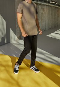 T-shirt olive vert à manches courtes, pantalon noir slim, et baskets noir et blanc avec des accents en daim, debout sur un sol jaune.