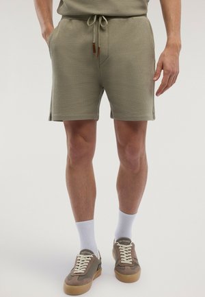 Uomo che indossa pantaloncini leggeri color oliva chiaro con coulisse, calzini bianchi e scarpe casual taupe con suole in gomma.