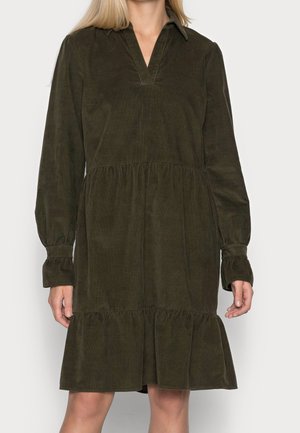 Frau trägt ein dunkelolivgrünes, langärmliges Cordkleid mit Kragen, V-Ausschnitt, geraffter Taille und Rüschensaum.