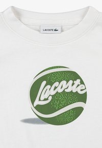 Λευκό βαμβακερό μπλουζάκι με κυκλικό πράσινο γραφικό με υφασμάτινο σχέδιο και τη λέξη "Lacoste" σε λευκά γράμματα στο κέντρο.