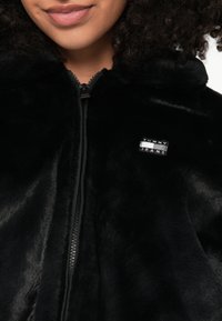 Γυναίκα που φοράει μαύρο τζάκετ με φερμουάρ και μικρό λογότυπο Tommy Jeans στο αριστερό στήθος, με το πρόσωπό της να φαίνεται με χαμόγελο.