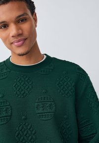 Ciemnozielony sweter z teksturowanymi wzorami świątecznych ozdób. Okrągły dekolt z prążkowanym wykończeniem. Miękki materiał z wypukłym wzorem.