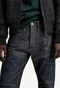 Donkerblauwe spijkerbroek met gele stiksels, voorzien van een knoopsluiting, schuine voorkruiken en een gladde textuur. De broek is gedeeltelijk bedekt door een zwarte bomberjack.