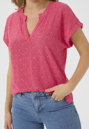 Mujer que lleva una camiseta rosa texturizada de manga corta con escote en V y vaqueros azules, con una mano en el bolsillo.