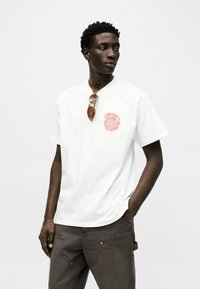 Άνδρας με λευκό T-shirt που φέρει κόκκινο λογότυπο σπείρας και καφέ παντελόνι, με γυαλιά ηλίου κρεμασμένα στον γιακά, στέκεται μπροστά σε απλό ανοιχτό φόντο.